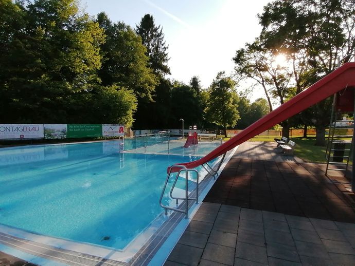 Freibad mit klarem, blauem Wasser, roter Rutsche und Bäumen im Hintergrund