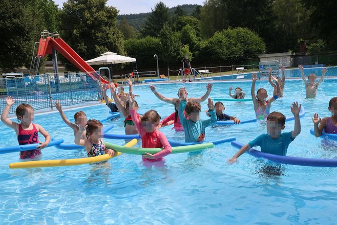 Kinder treiben lächelnd und mit erhobenen Armen in einem Freibad auf bunten Ringen in einem Pool.