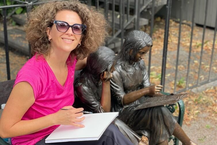 Eine Frau in einem rosa Hemd lächelt neben einer sitzenden Bronzestatue und hält ein Notizbuch im Freien an einem Zaun.
