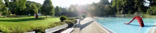 Ein Panoramabild des Freibades Oberbieber. Eine Wiese links, eine rote Rutsche in der Mitte und mehrere Schwimmbecken rechts.