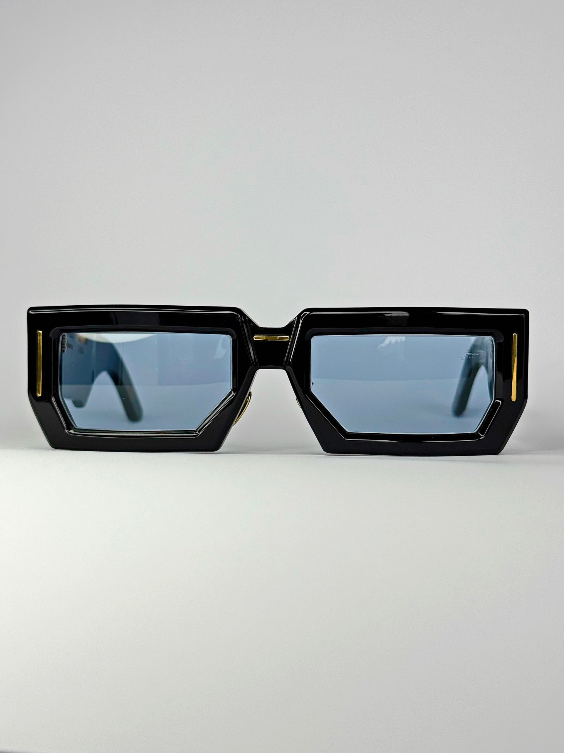 Schwarze, eckige Sonnenbrille mit blauen Gläsern vor weißem Hintergrund.