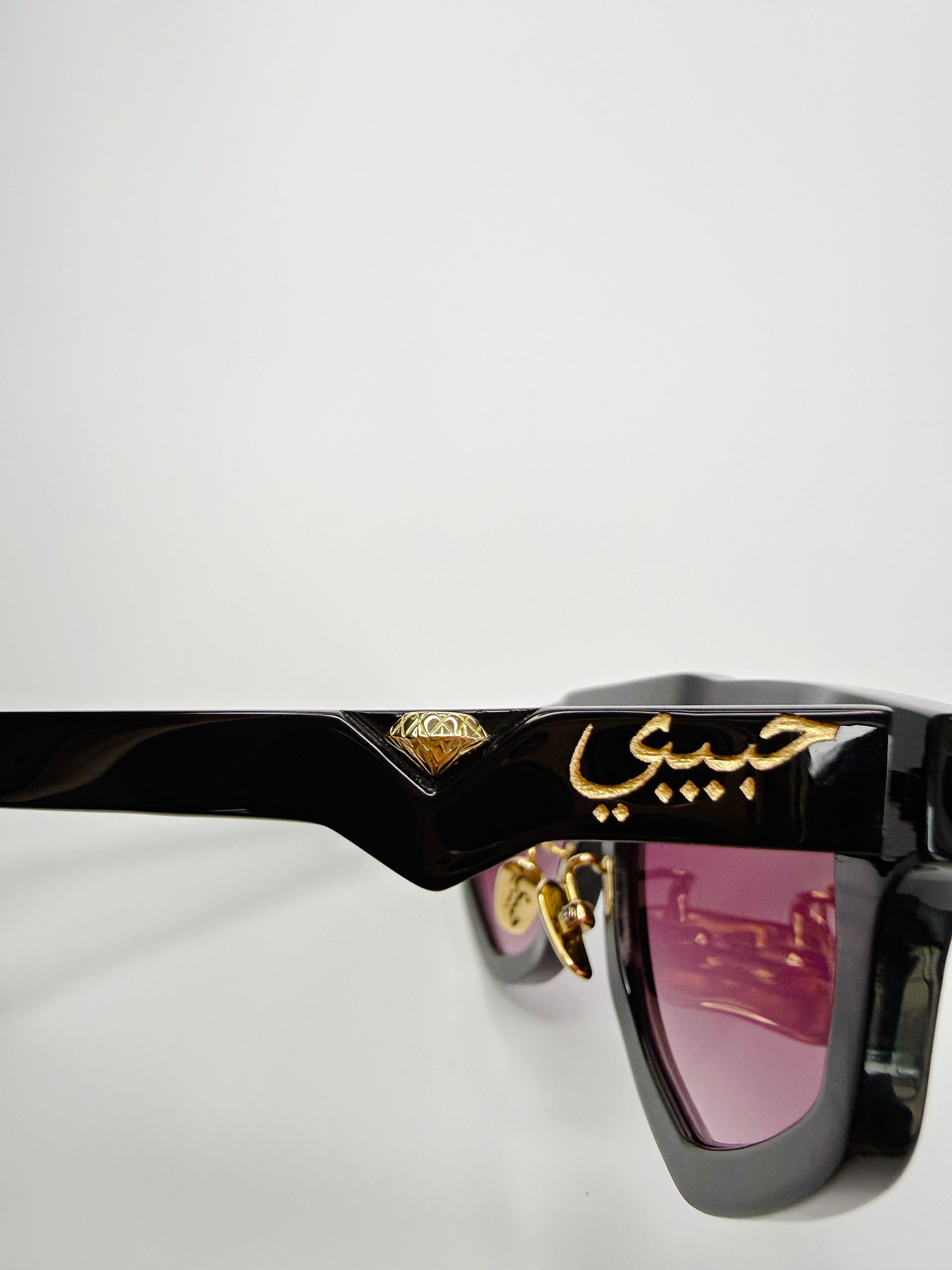 Schwarze Sonnenbrille mit goldener arabischer Schrift „habibi“ auf dem Rahmen, rosa Gläser.