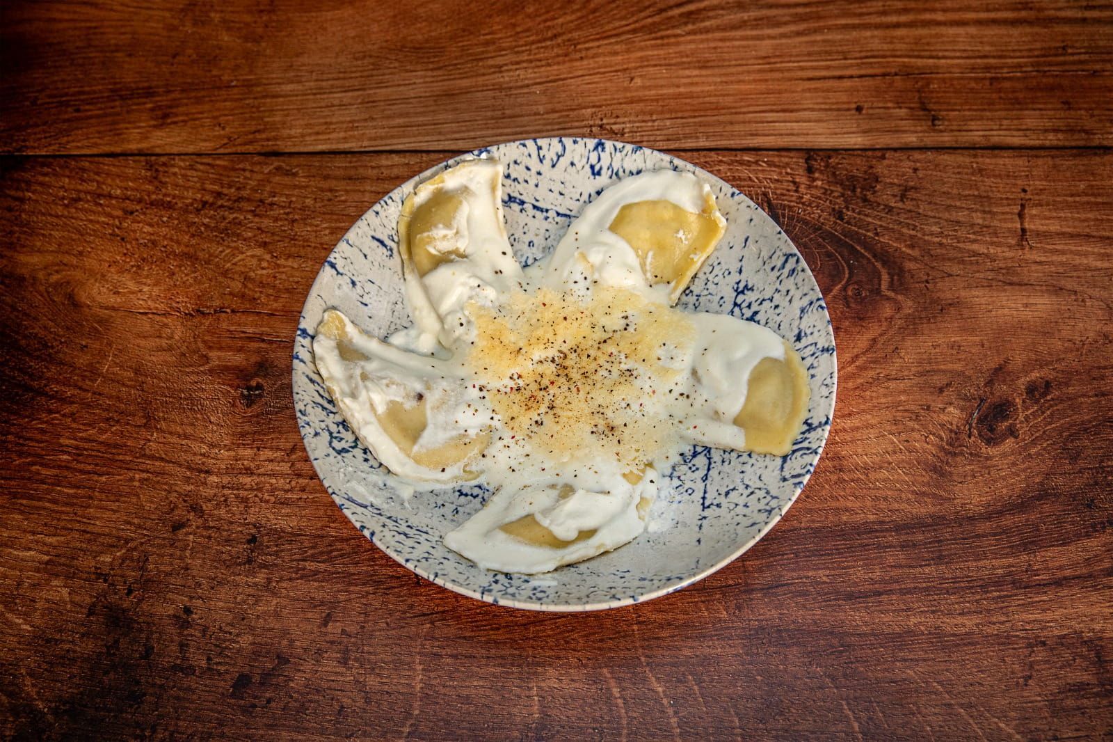 Pierogies en un plato blanco con motas azules, cubiertos con salsa de crema y pimienta, sobre una mesa de madera.