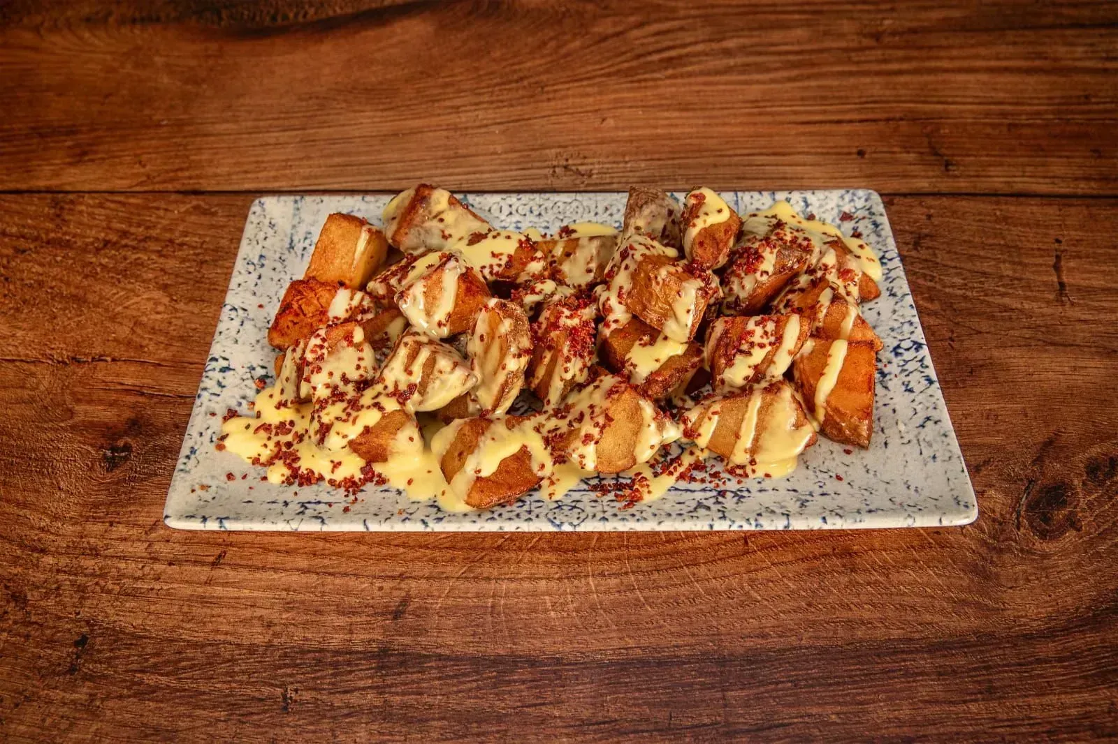 Papas fritas con salsa amarilla y chispitas rojas sobre un plato con estampado blanco encima de una mesa de madera.