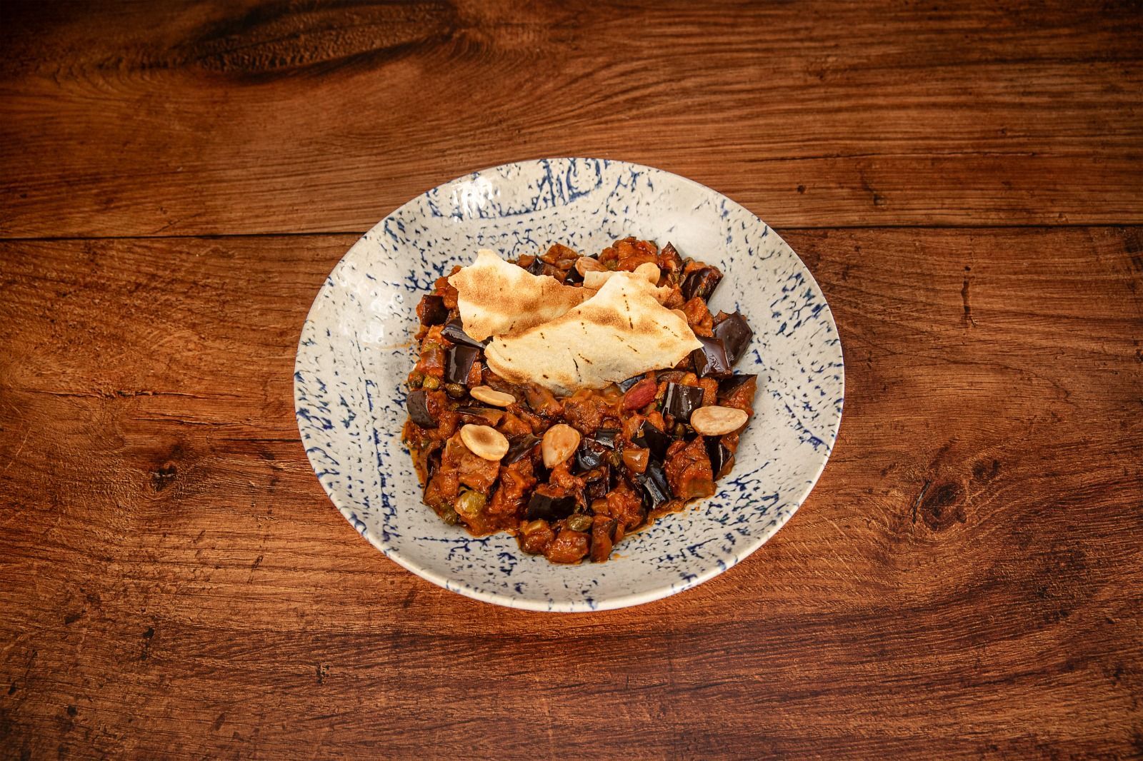 Plato de guiso de berenjenas con nueces y crema en un plato con borde azul, colocado sobre una superficie de madera.