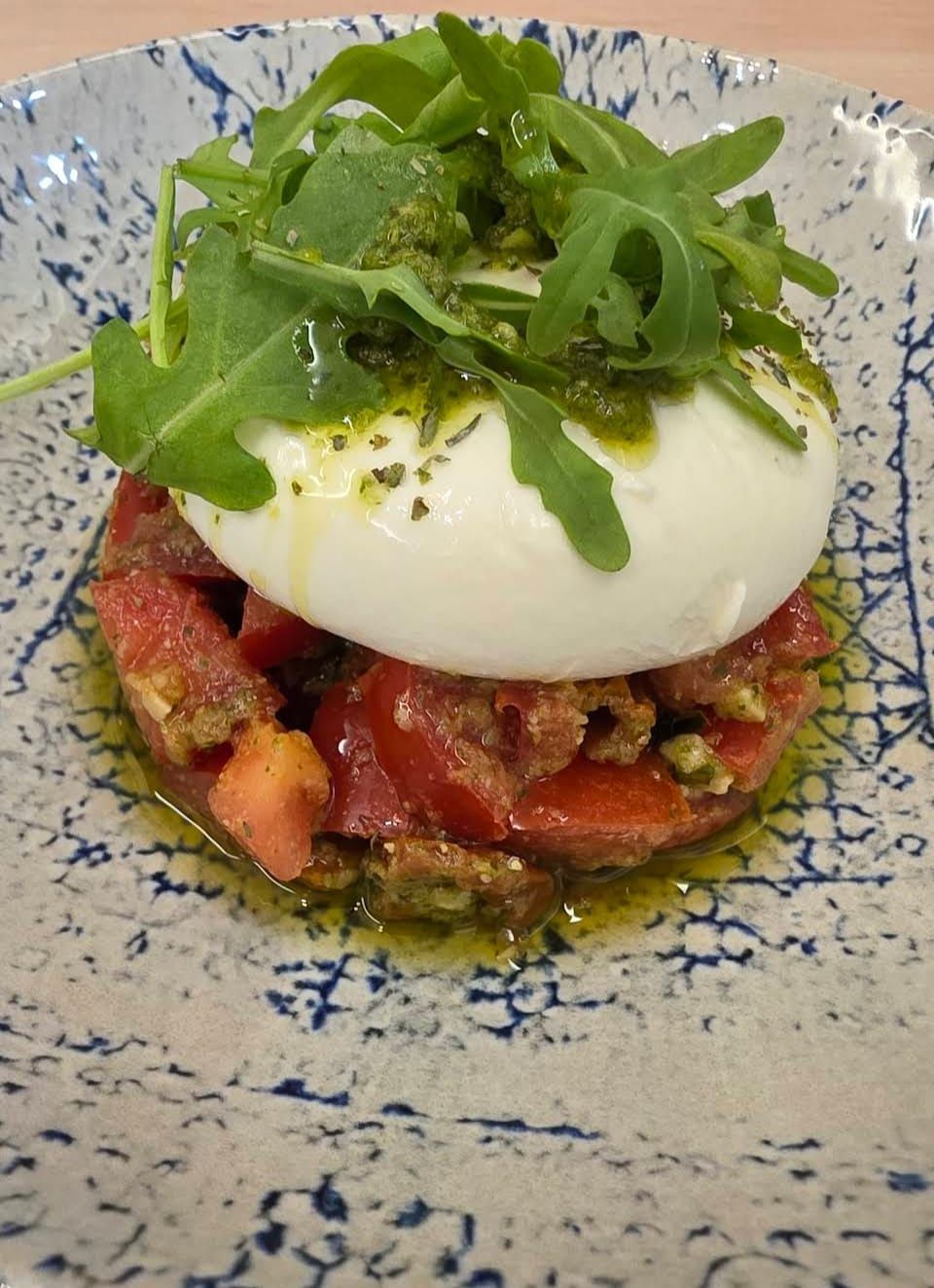 Ensalada de burrata y rúcula sobre rodajas de tomate, rociada con pesto, en plato estampado.