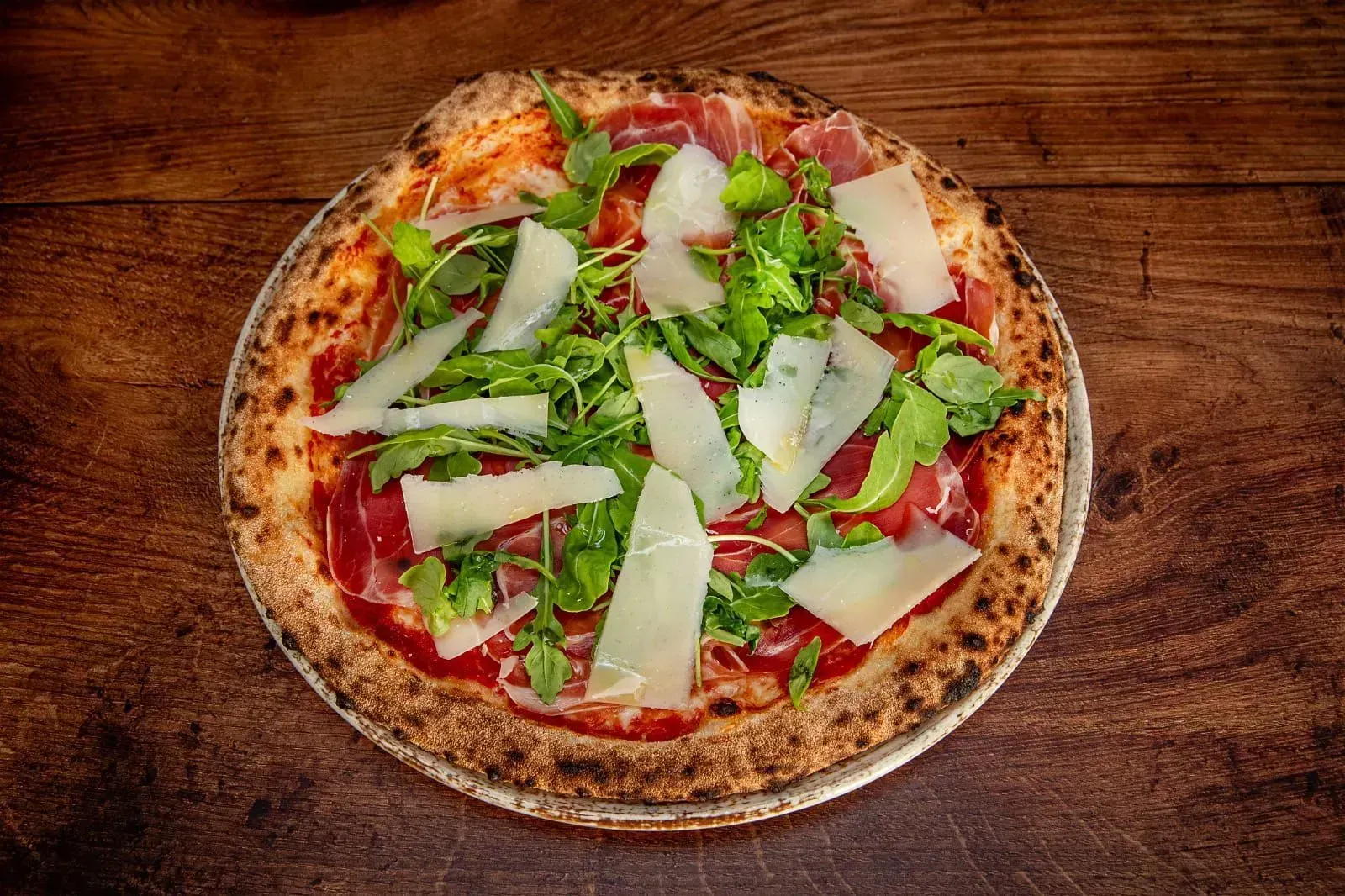 Pizza cubierta con rúcula, parmesano rallado y prosciutto sobre una mesa de madera.