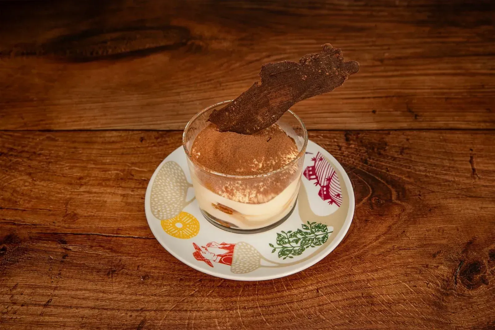Postre de tiramisú en copa, cubierto con cacao en polvo y decoración de chocolate, sobre platillo estampado, sobre madera.