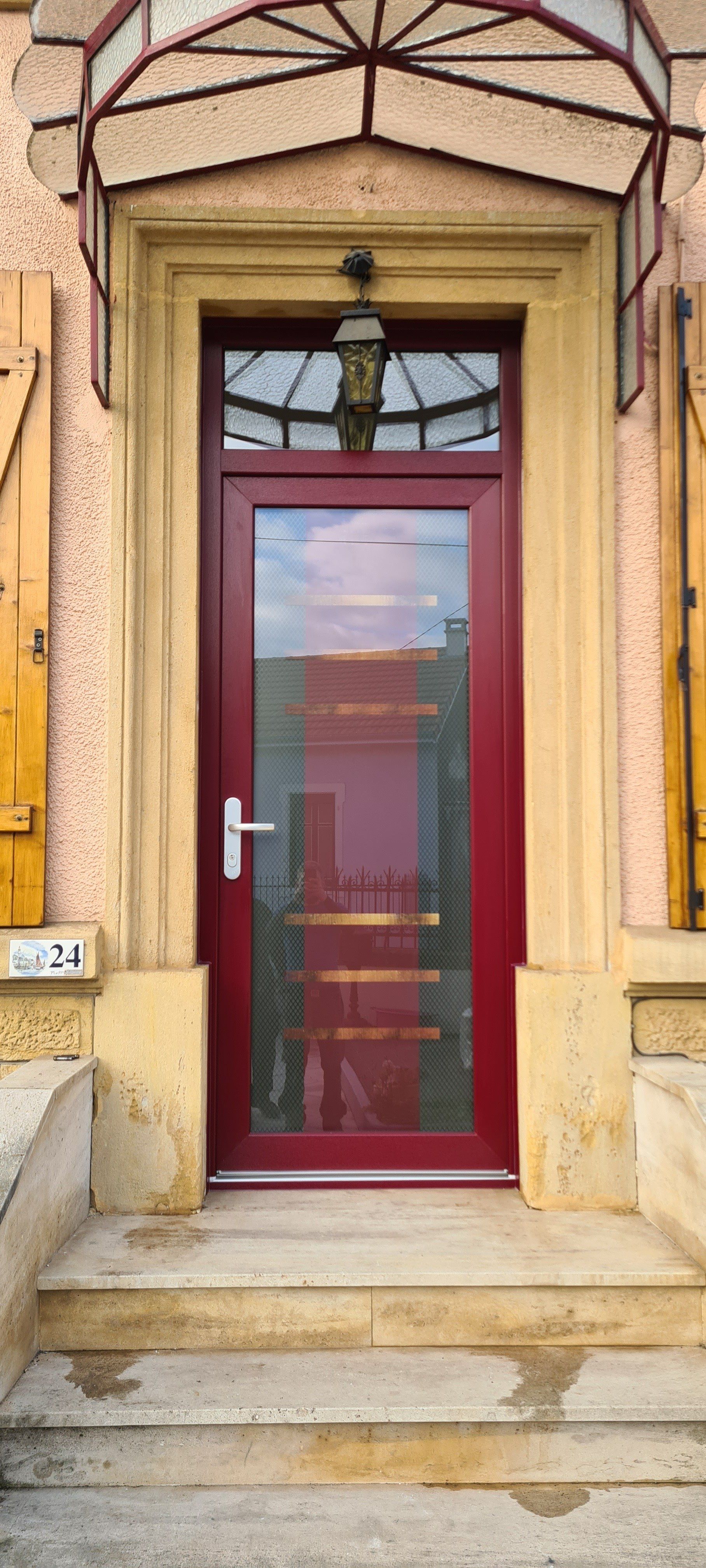 Porte d'entrée en bois de couleur rouge avec de la vitre incrustée sous un carport en verre et fer forgé.