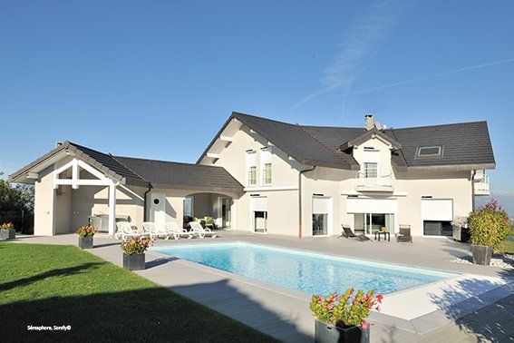 Grande maison avec crépit blanc et toiture de tuile noire, avec piscine et volet roulant moitié fermée sur les menuiseries