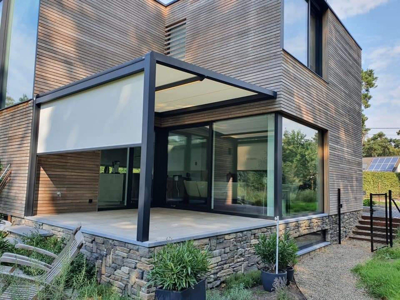 Pergola noire en alu avec occultant devant une maison moderne avec grande vitre qui fait l'angle de la maison
