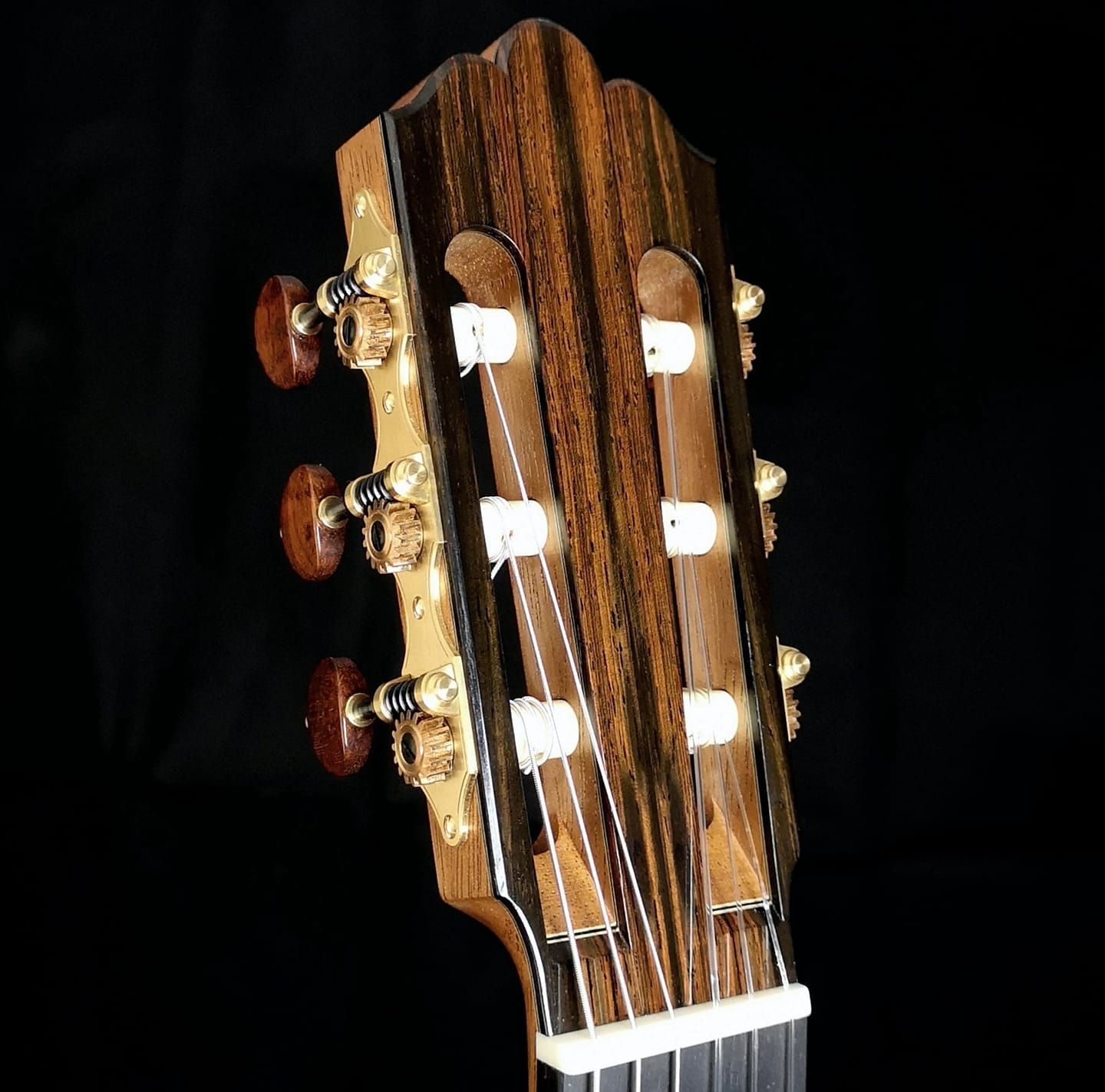 Clavijero de una guitarra clásica con veta de madera oscura y clavijas de afinación doradas sobre un fondo negro.