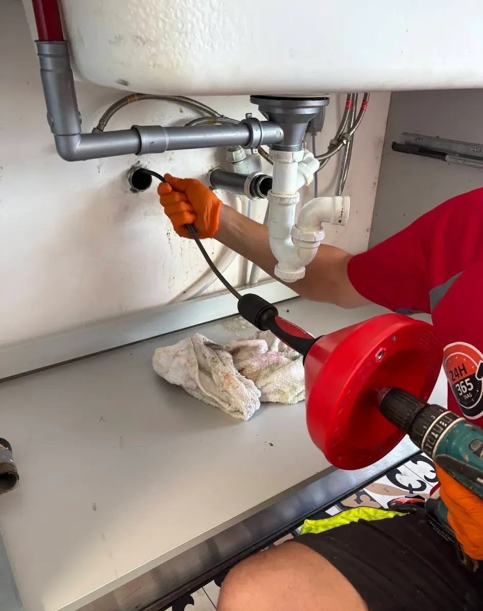 Persona destapando el desagüe de un fregadero de cocina con una herramienta desatascadora motorizada.