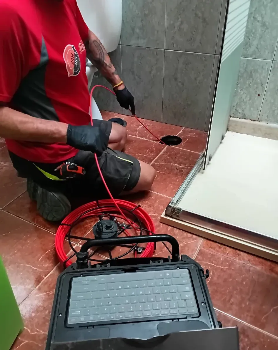 Un fontanero inspecciona un desagüe con una cámara en un baño. Camisa roja y negra, azulejos grises y un portátil.