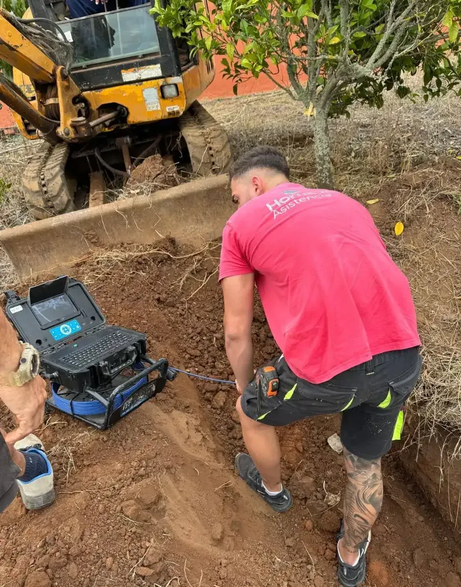 Hombre con camisa roja cerca de una zanja con equipo, una pequeña excavadora al fondo.