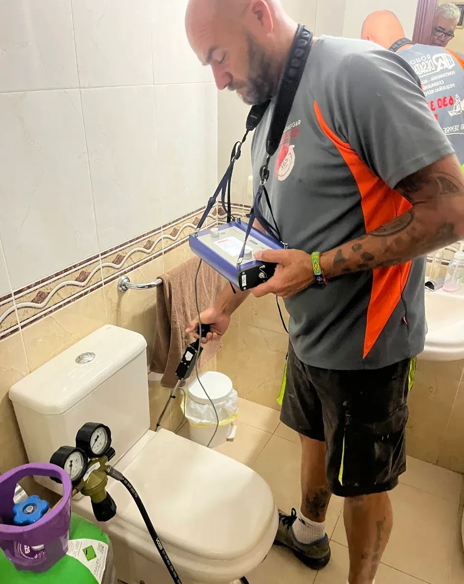 Hombre en el baño probando el inodoro con equipo. Inodoro blanco, tanque de gas y paredes de azulejos.