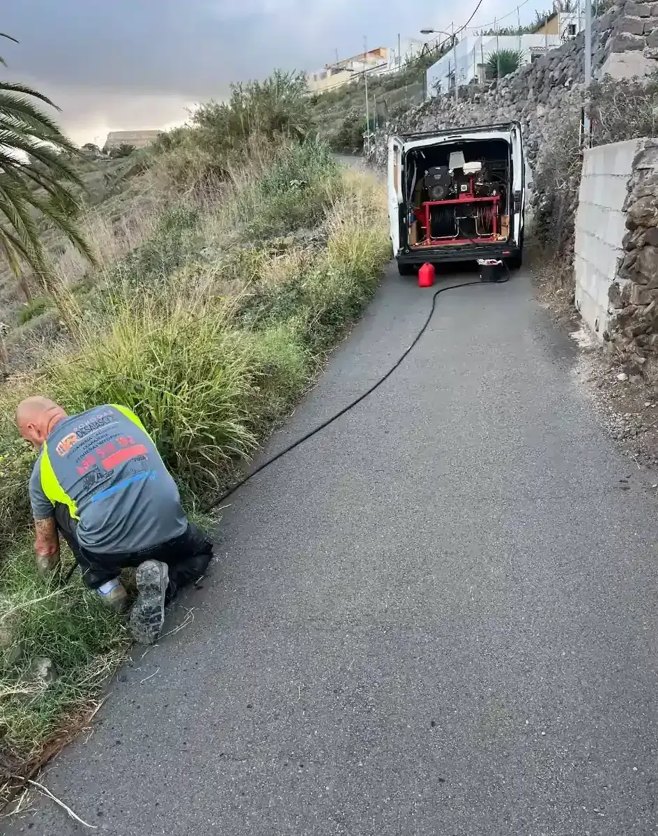 Hombre inspeccionando un cable en una carretera estrecha, con una camioneta de servicio estacionada detrás de él.