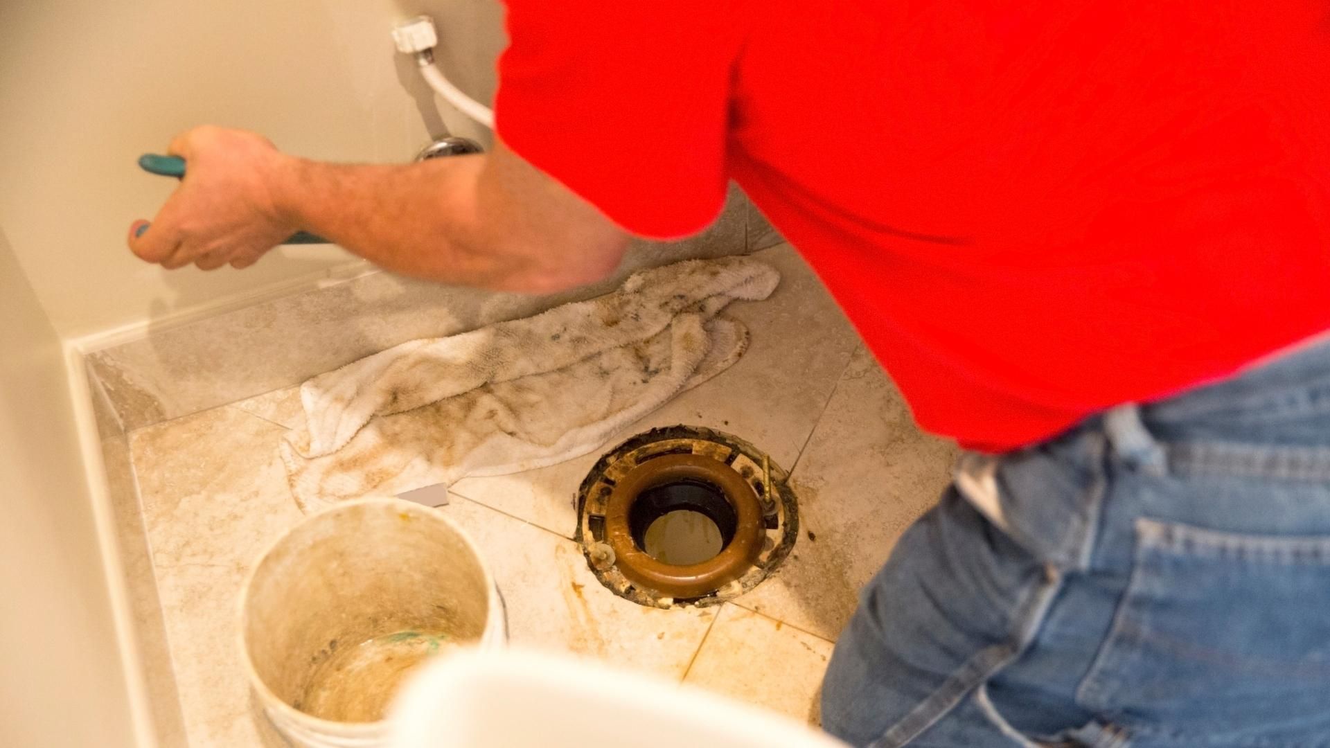Fontanero con camisa roja reparando un inodoro, sosteniendo una llave inglesa, de pie sobre un desagüe abierto.