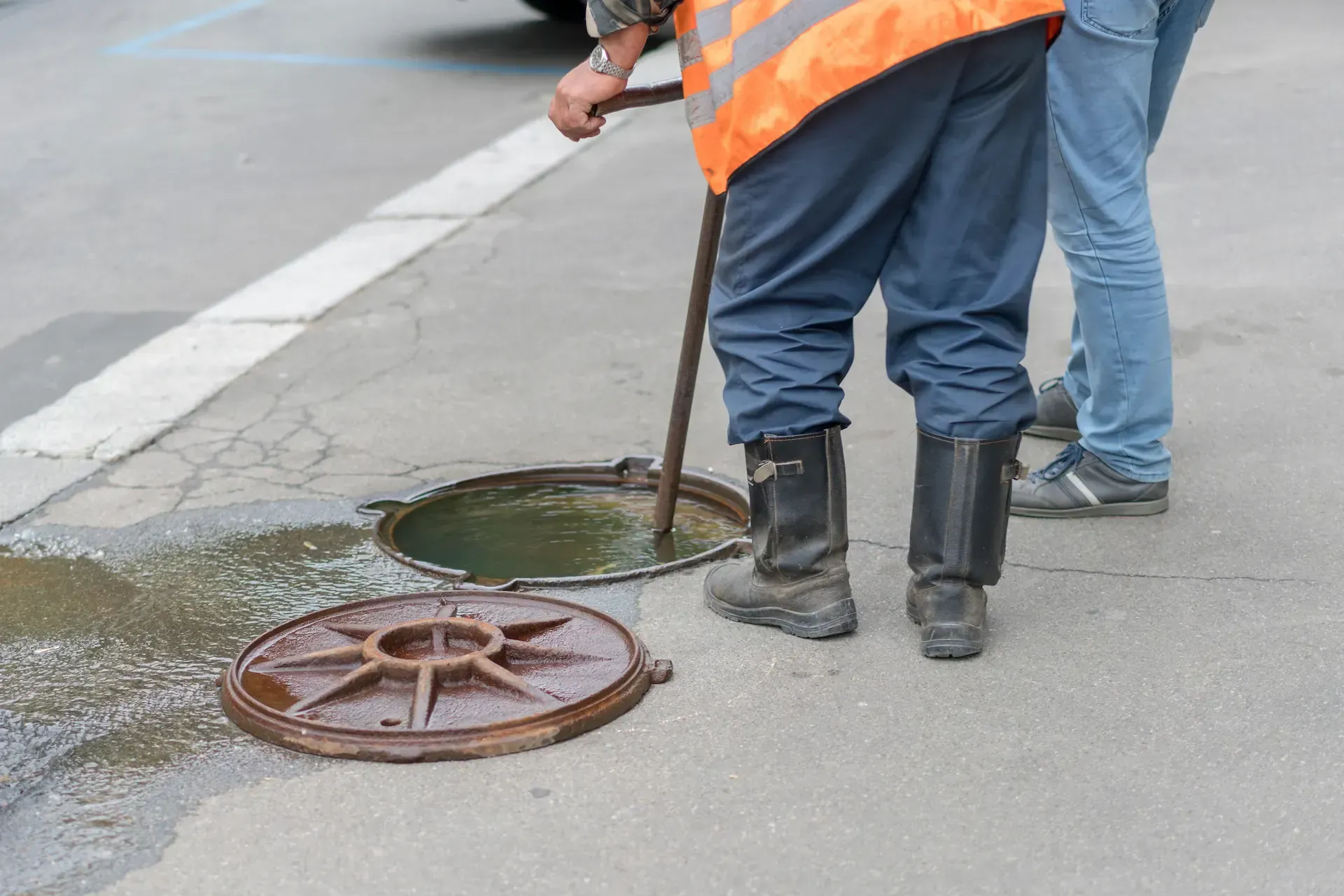 trabajadores abren una alcantarilla en una calle de la ciudad. Uno lleva un chaleco de seguridad y usa una herramienta.