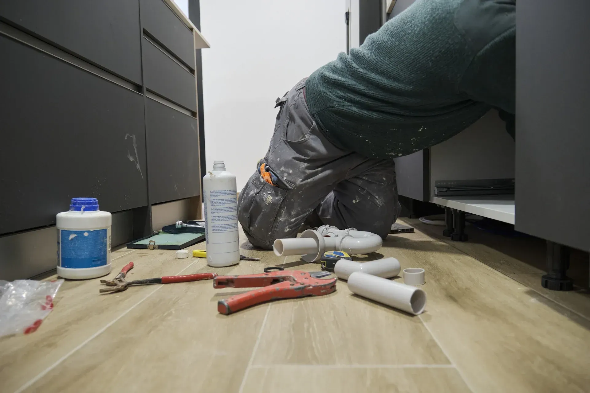 Fontanero trabajando en tuberías debajo de un armario oscuro en una cocina, herramientas y suministros en el suelo.