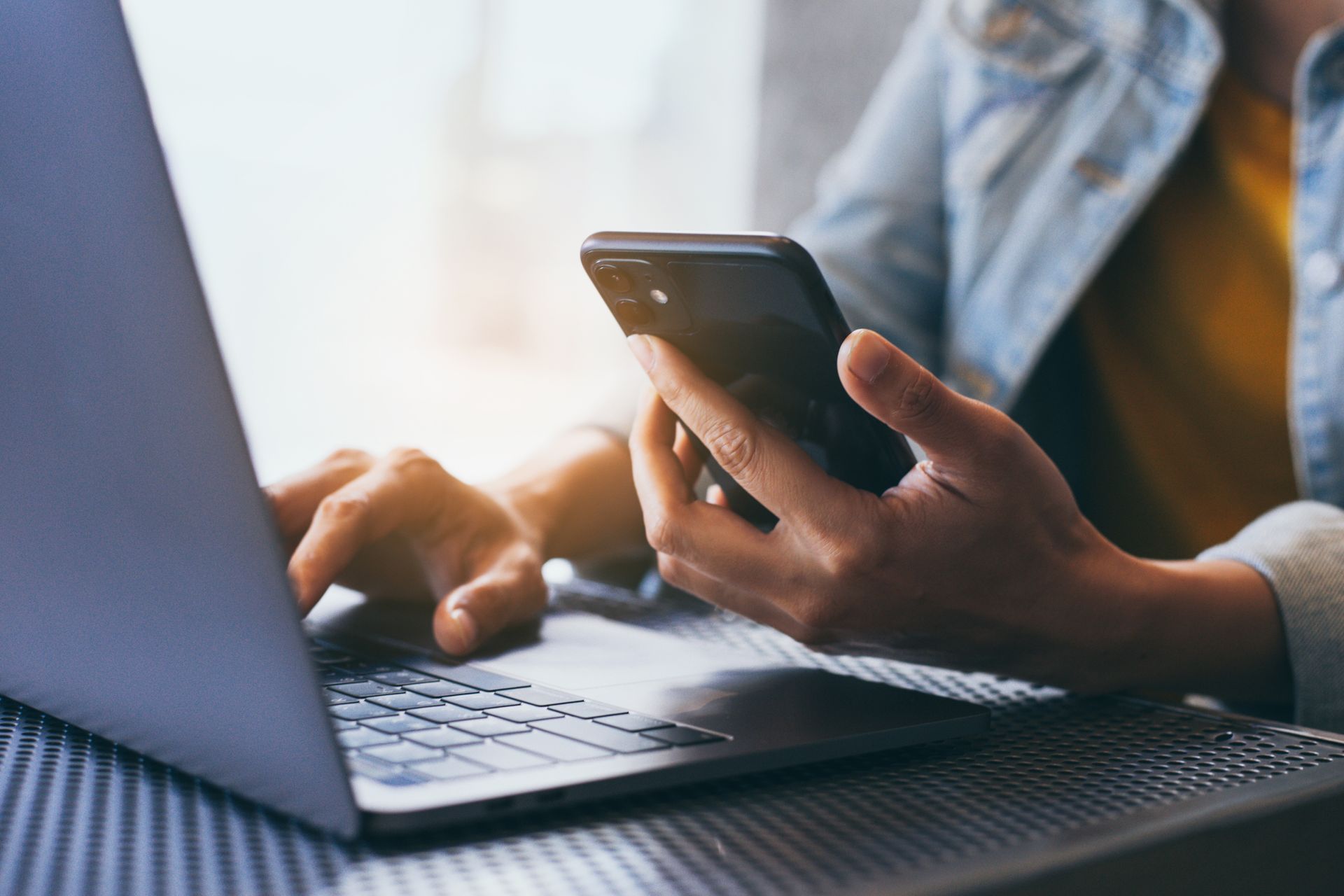 Des mains manipulant un ordinateur portable et un smartphone à un bureau, avec une chemise bleue en arrière-plan.