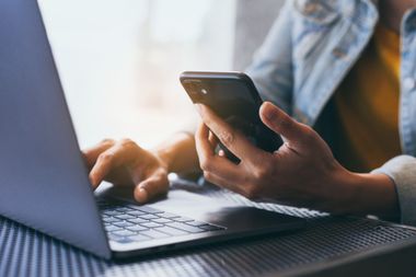 Des mains manipulant un ordinateur portable et un smartphone à un bureau, avec une chemise bleue en arrière-plan.