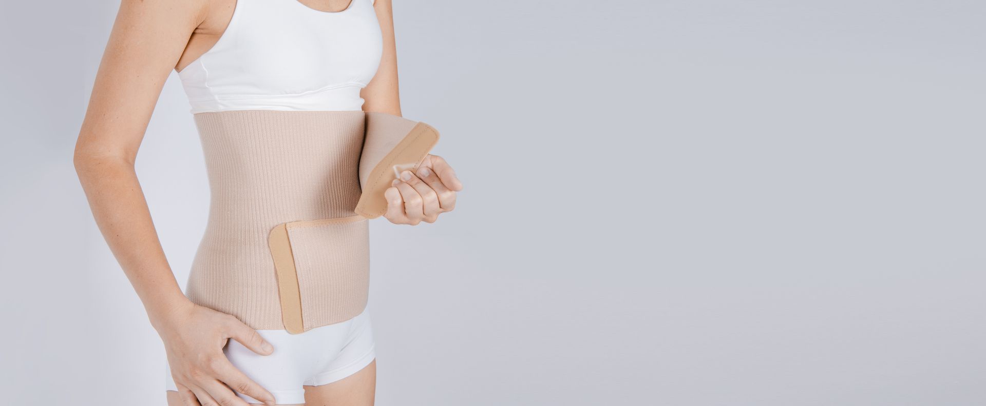 Femme portant une ceinture abdominale beige. Elle est vêtue d'un sous-vêtement blanc et d'un haut blanc. Fond gris.