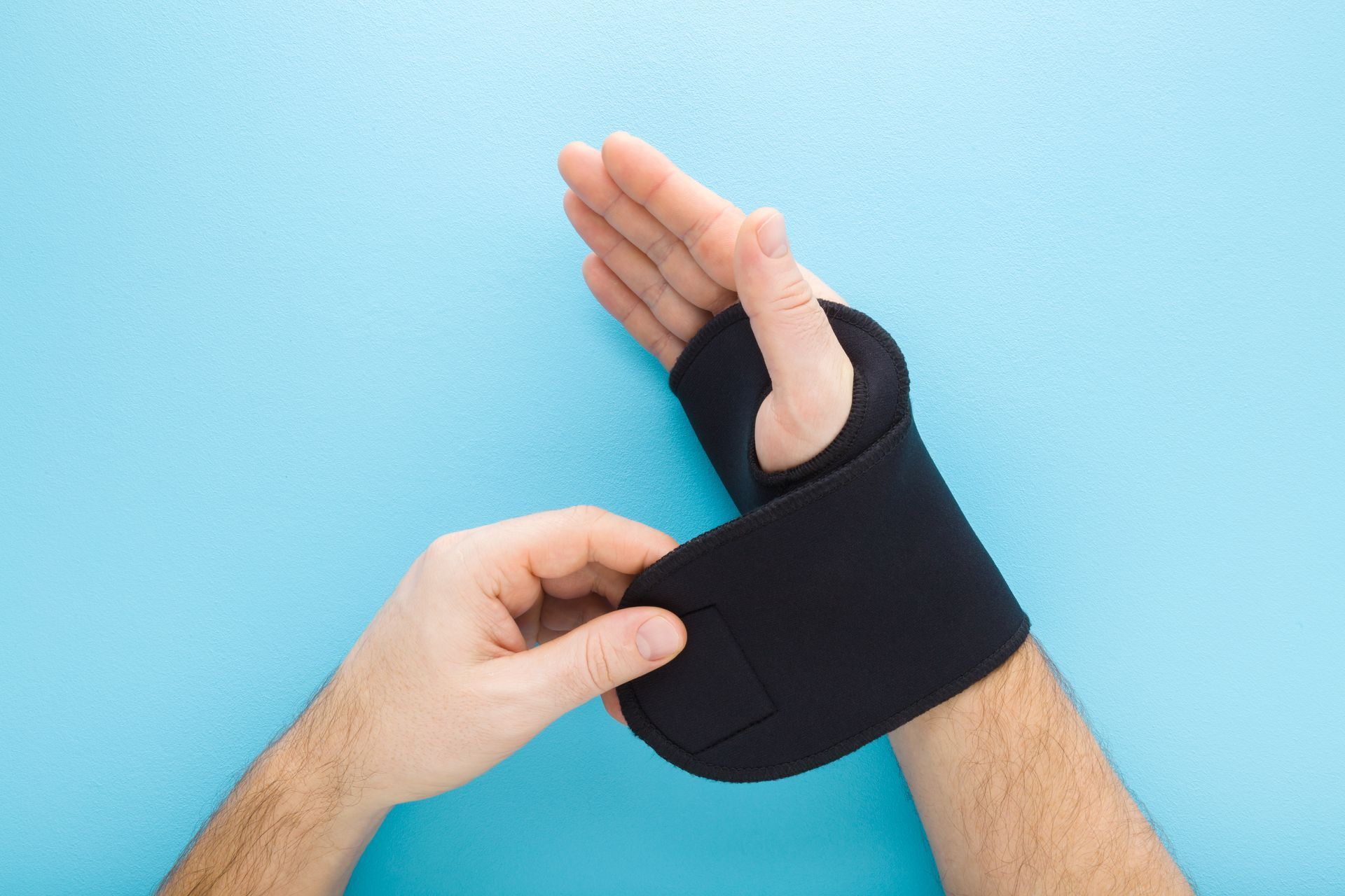 Main d'une personne portant une attelle de poignet noire, l'autre main ajustant la sangle, sur fond bleu.