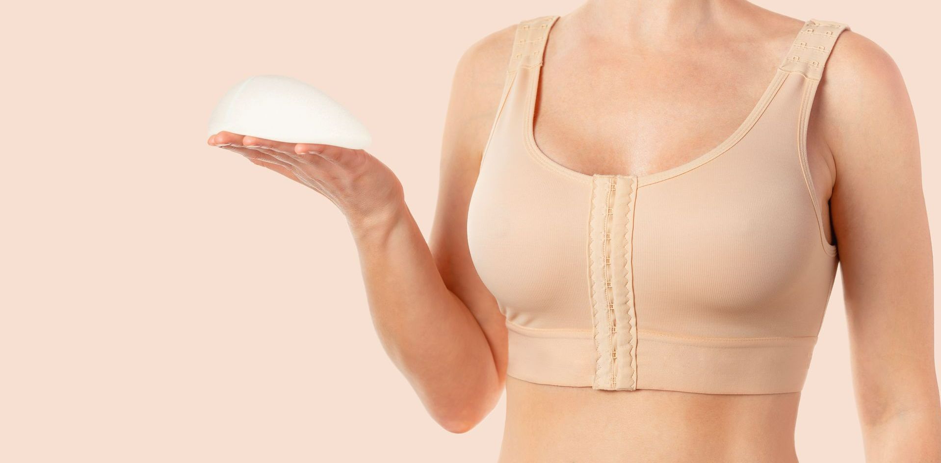 Femme portant un soutien-gorge de contention tenant un implant mammaire. Fond pêche.