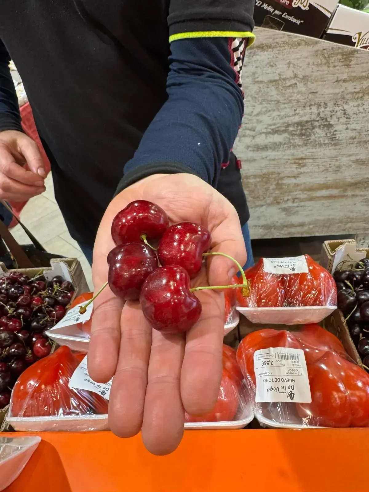 Una mano sostiene un racimo de cerezas rojas en un puesto de mercado con fruta envasada al fondo.