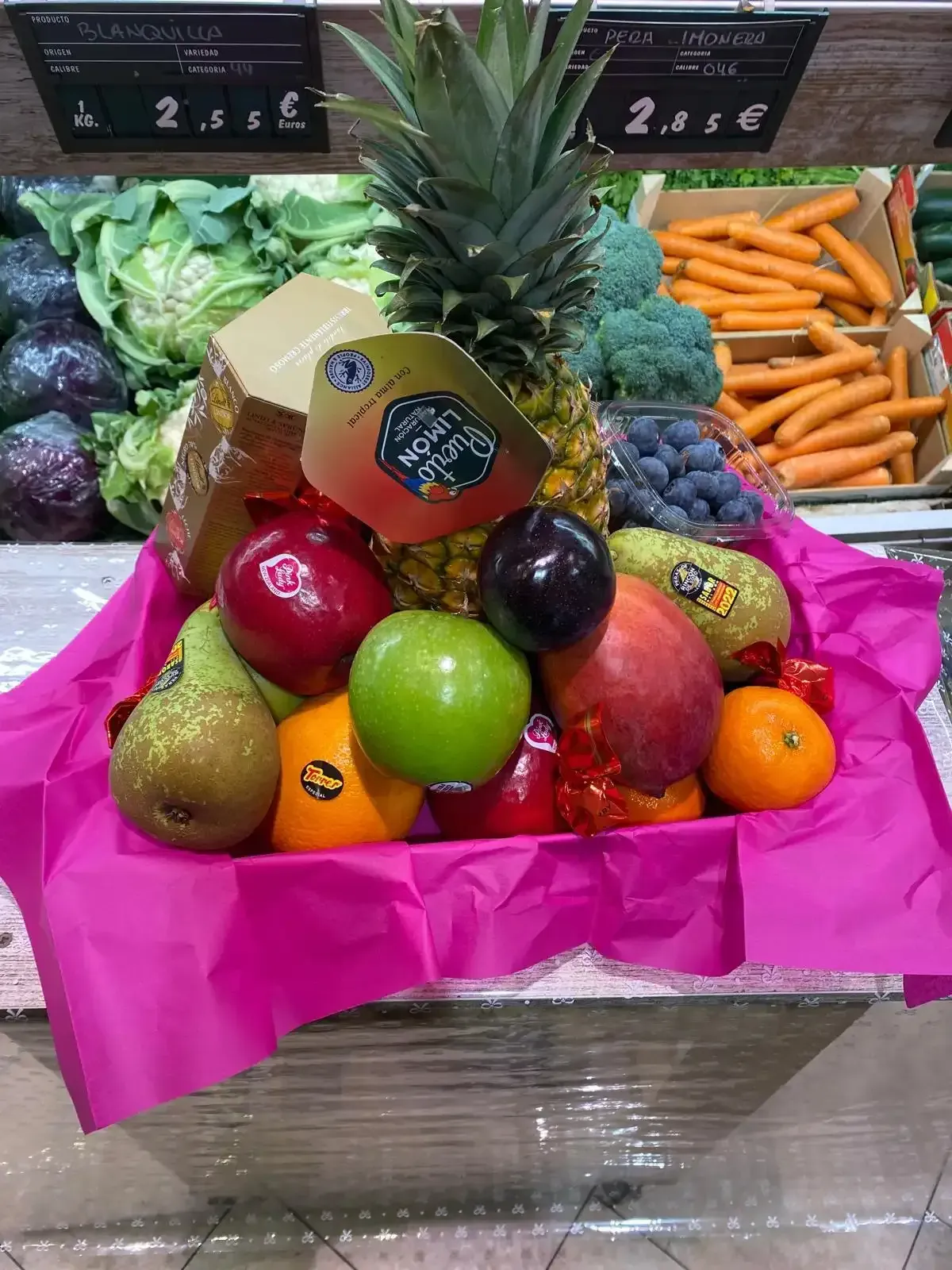 Cesta de frutas con piña, manzanas, naranjas, uvas y bayas en un puesto de mercado con verduras al fondo.