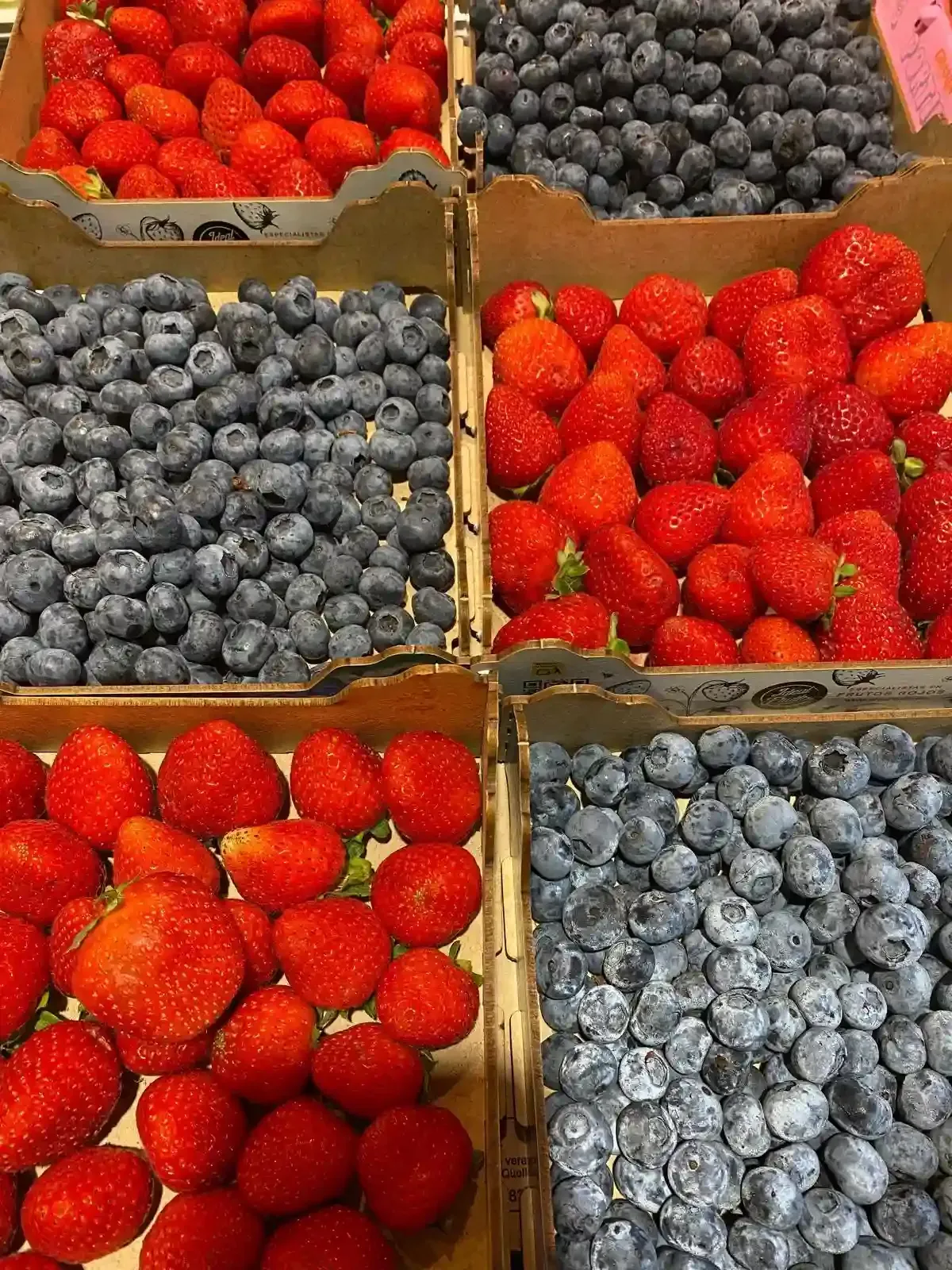 Seis cajas de cartón llenas de fresas y arándanos frescos en un puesto del mercado.