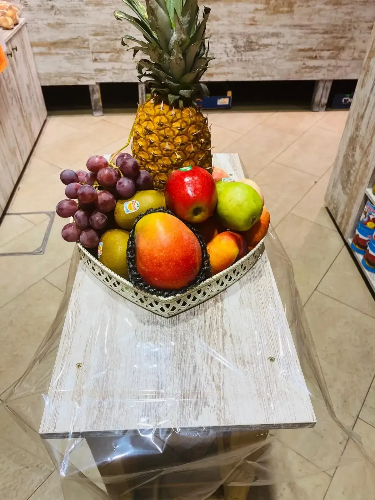 Cesta de frutas con piña, uvas, mango, manzanas y peras sobre una mesa blanca.