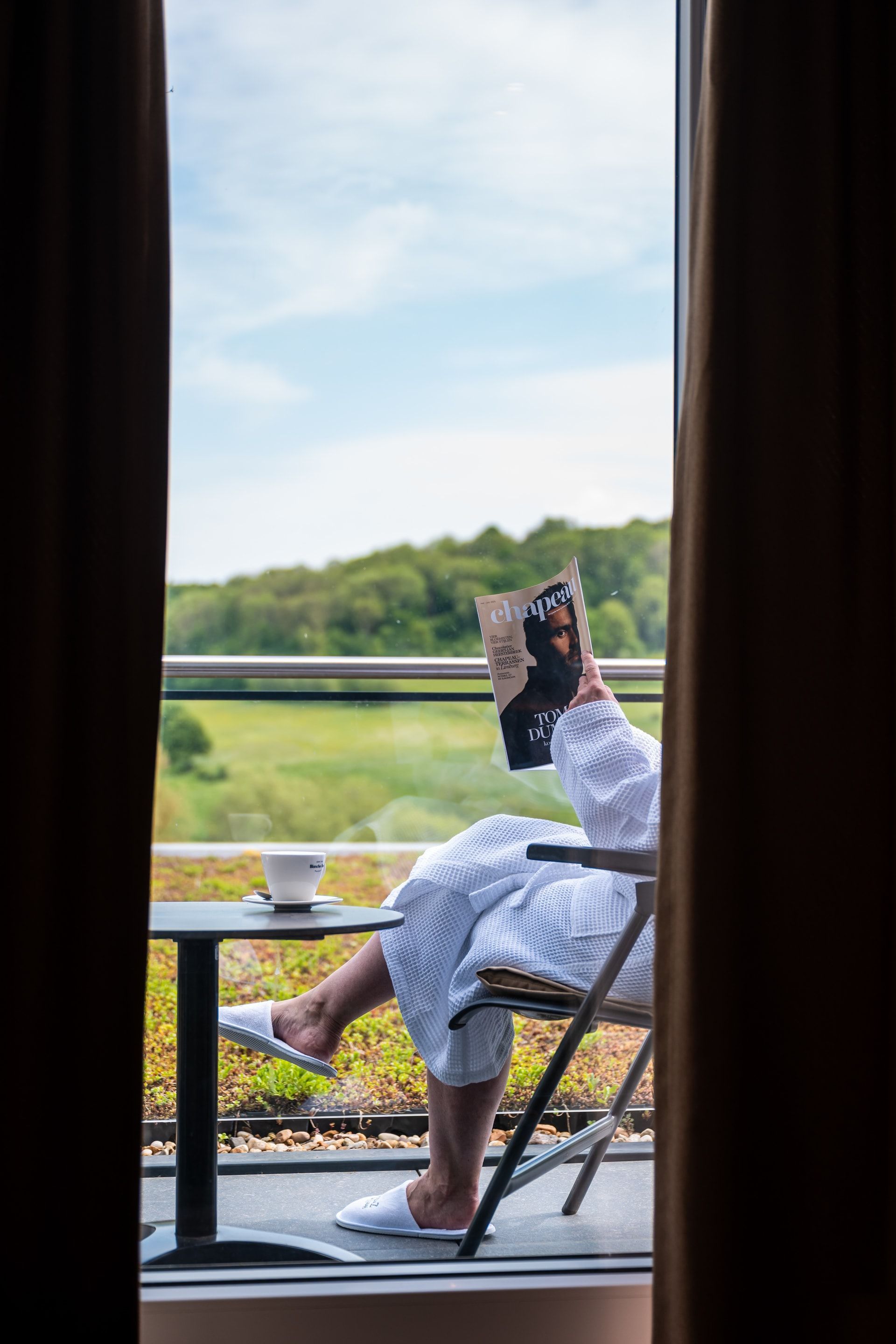 Een vrouw in een badjas zit op een balkon en leest een boek.