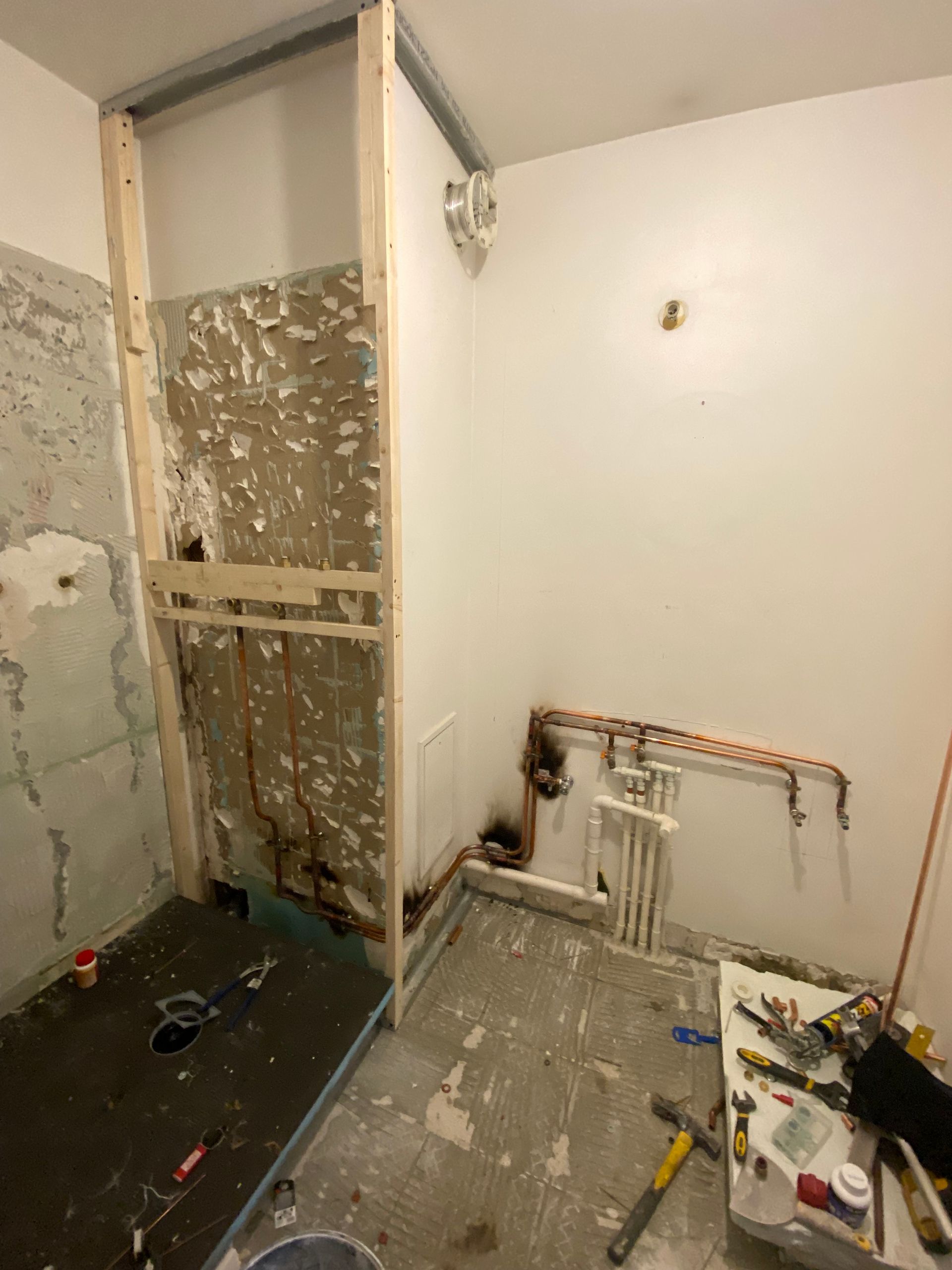 Salle de bains partiellement démolie, murs apparents, débris au sol et tuyauterie visible.