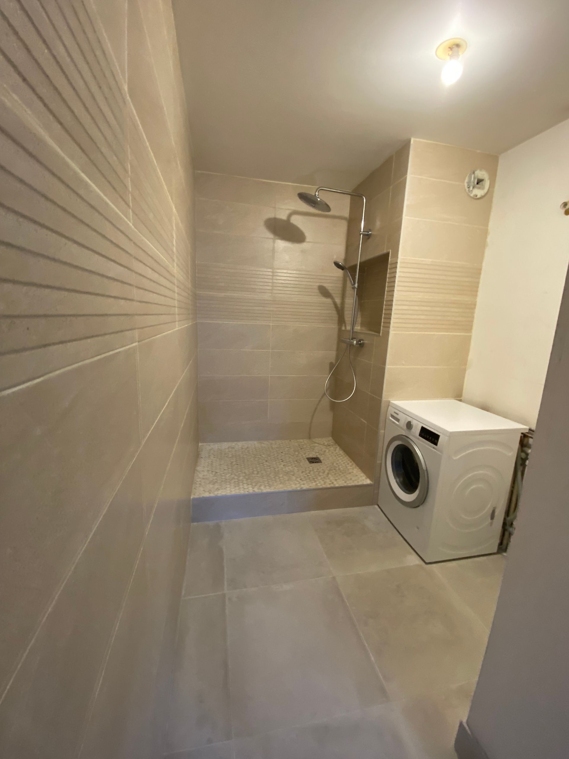Salle de bains étroite carrelée de beige avec douche, lave-linge et éclairage au plafond