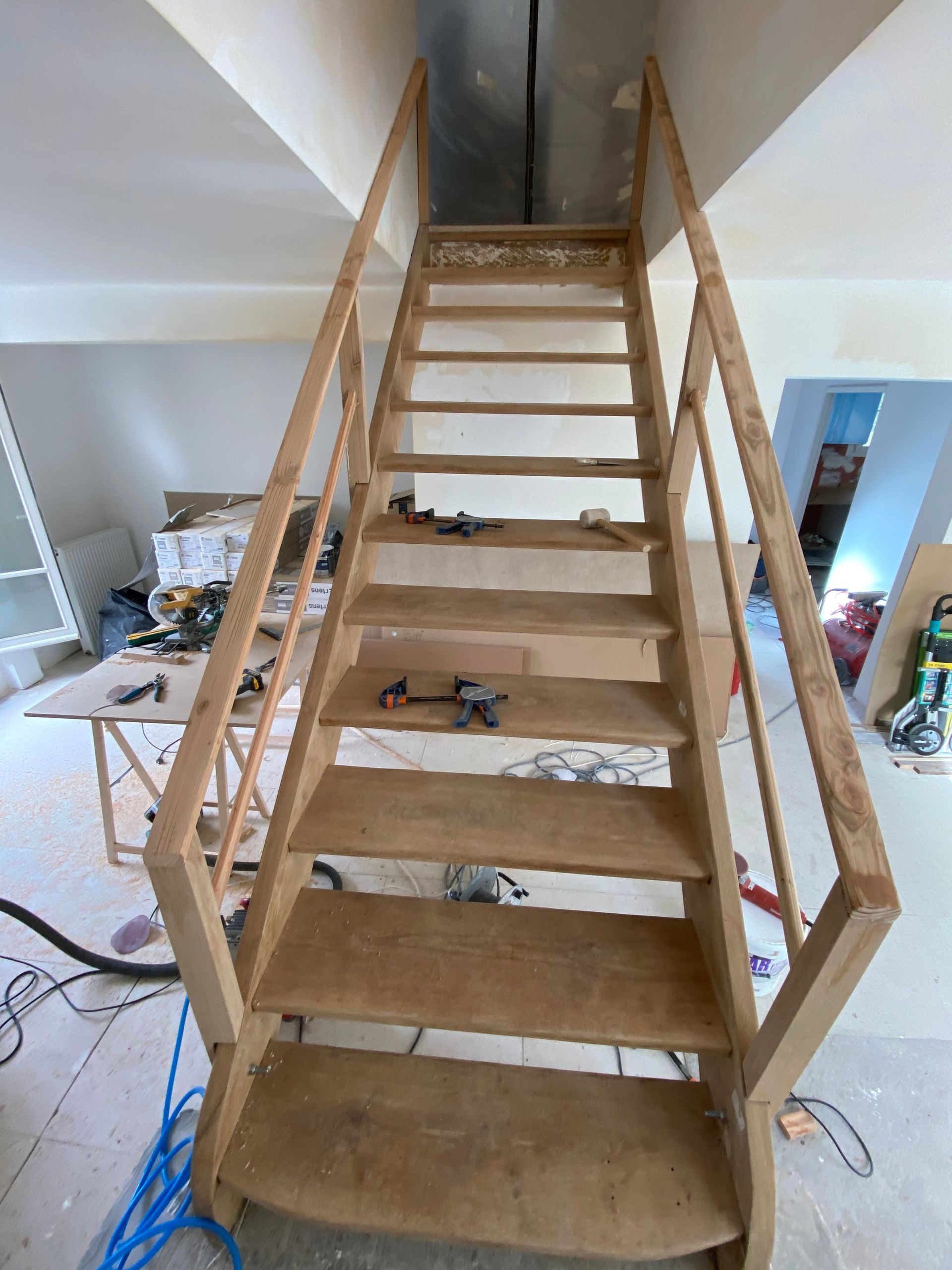 Un escalier en bois est en construction dans une pièce , avec des outils et des câbles visibles en dessous.