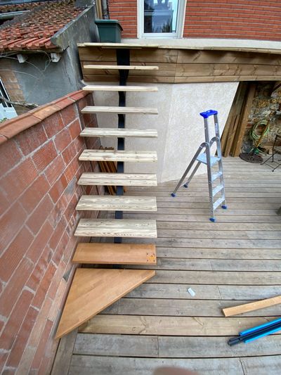 Escalier en bois en construction le long d'un mur de briques, avec une échelle sur une terrasse en bois.