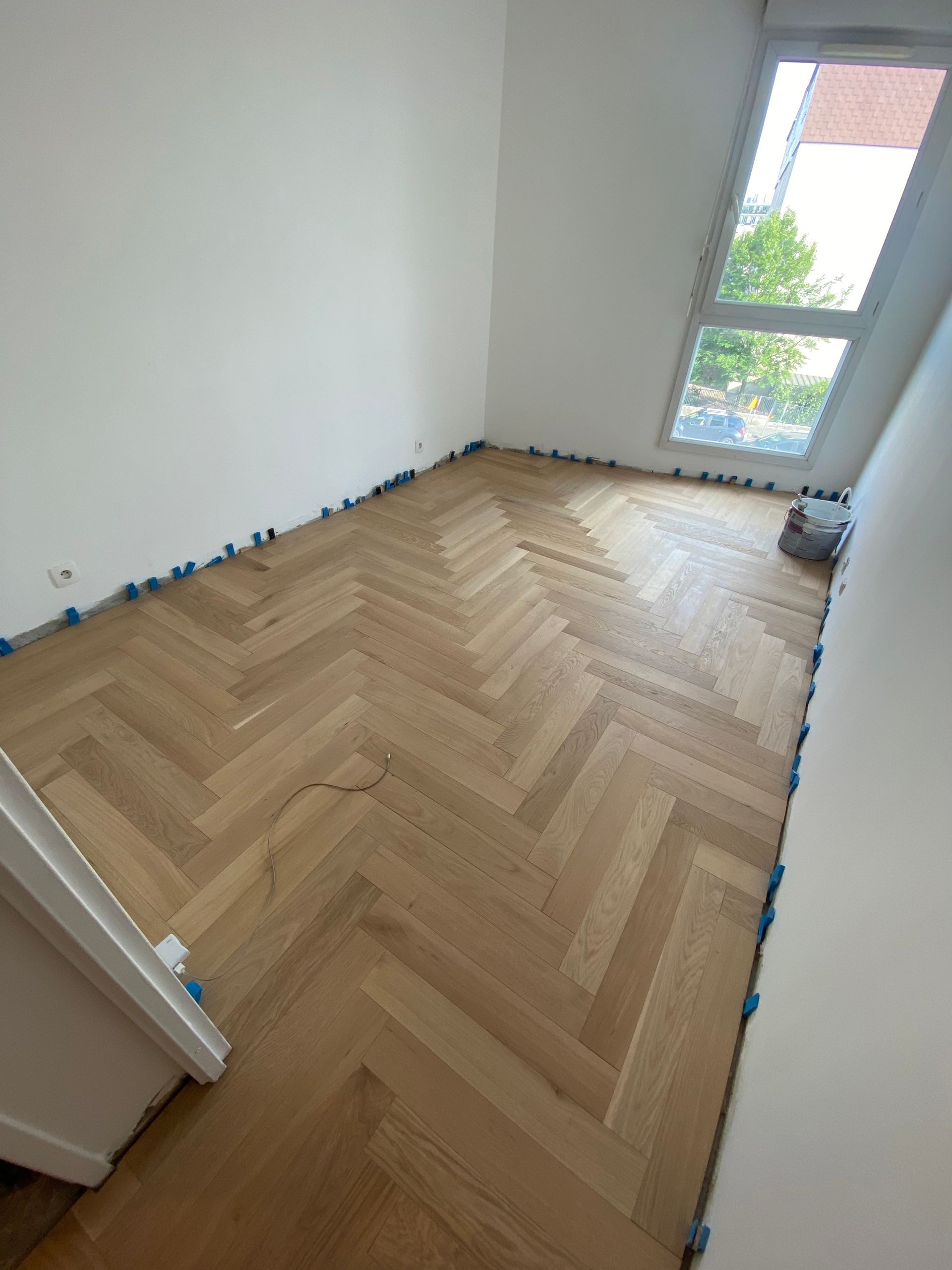 Pièce vide avec parquet à chevrons, murs blancs et une fenêtre.