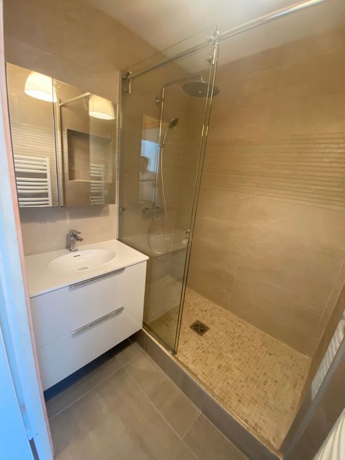 Salle de bains moderne avec meuble-lavabo blanc, miroir et douche à l'italienne en verre carrelée de beige.