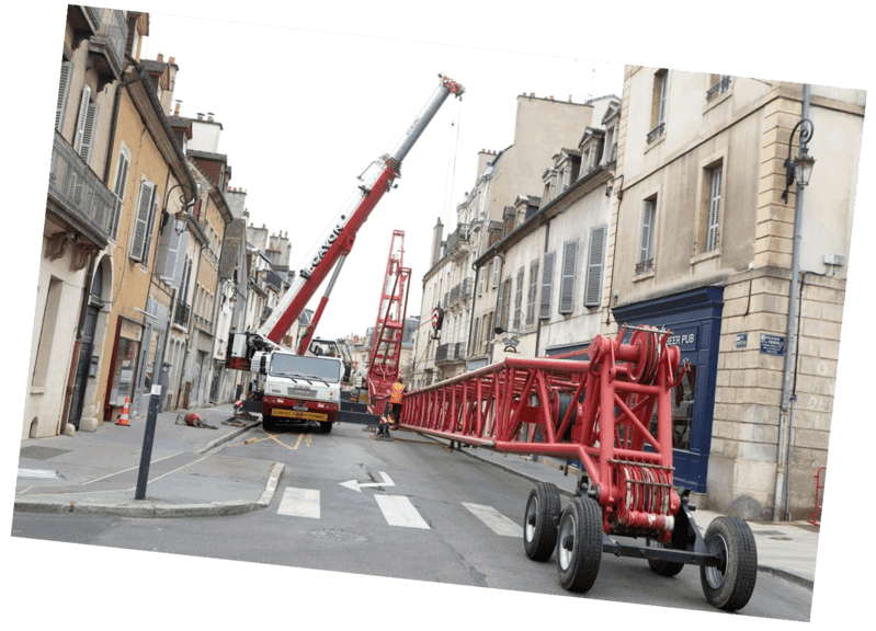 Une grue de chantier rouge est en cours d'assemblage dans une rue étroite de la ville, près de bâtiments beiges.