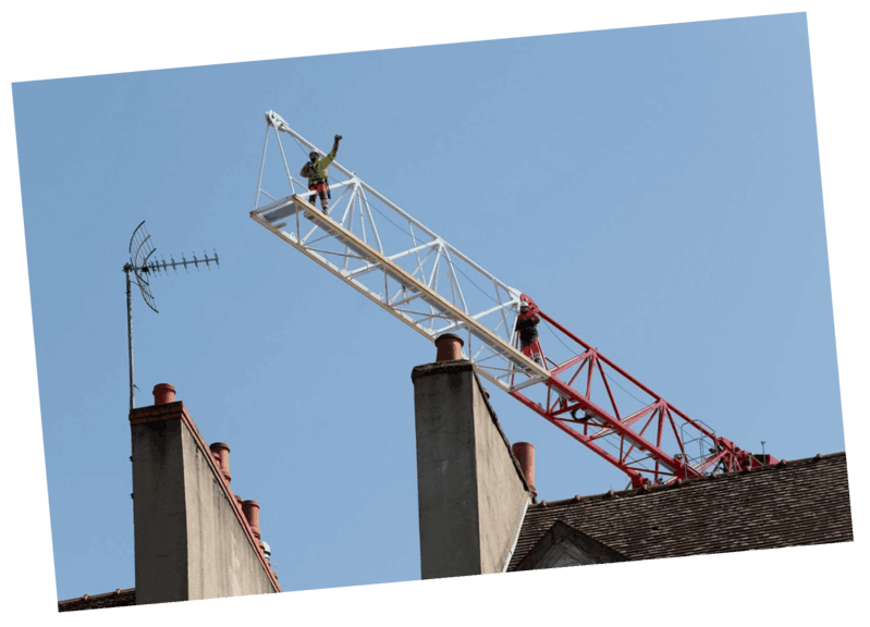 Des ouvriers travaillent sur le bras d'une grue au sommet d'un immeuble ; un bras blanc et rouge se déploie dans le ciel bleu.