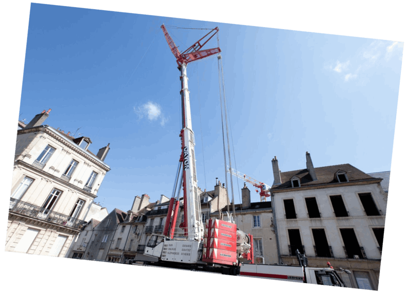 Une grande grue de chantier dans un cadre urbain, avec des bâtiments et un ciel bleu en arrière-plan.