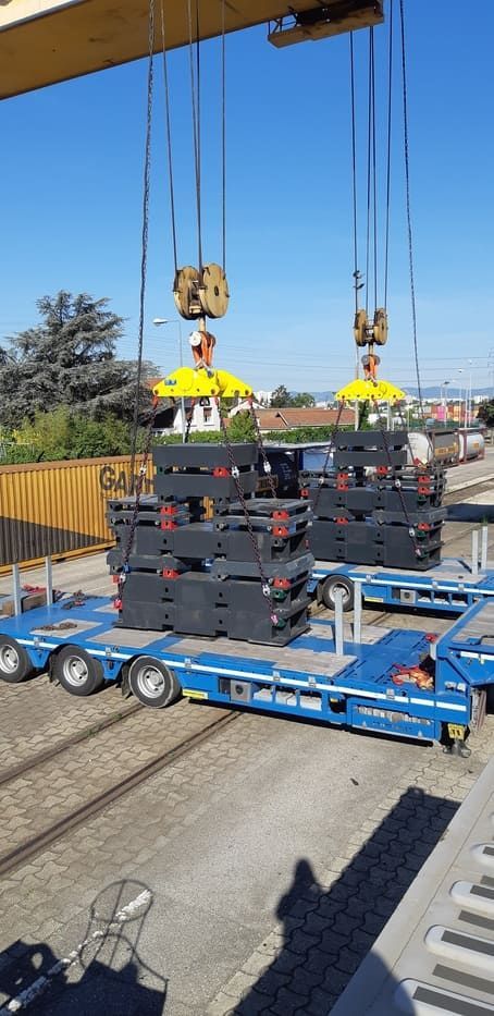 Des grues soulèvent des piles d'objets gris foncé sur des remorques bleues stationnées près des voies ferrées. Ciel bleu clair en arrière-plan.
