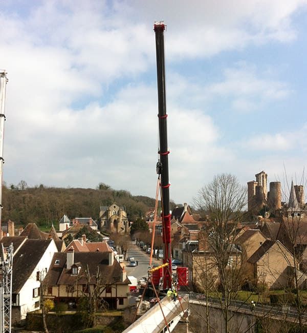 Une grande grue soulève des charges sur un pont en ville. Des bâtiments et des arbres entourent la grue, sous un ciel nuageux.