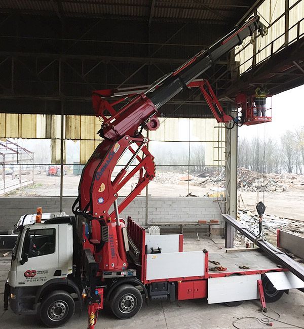Camion-grue rouge avec un ouvrier dans la nacelle levée, travaillant à l'intérieur d'un bâtiment.
