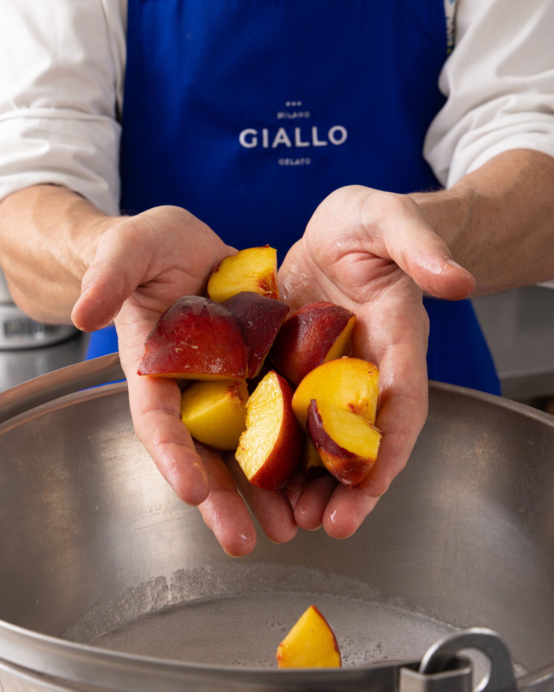 Mani che tengono pesche tagliate sopra una ciotola di metallo; grembiule blu con il logo 