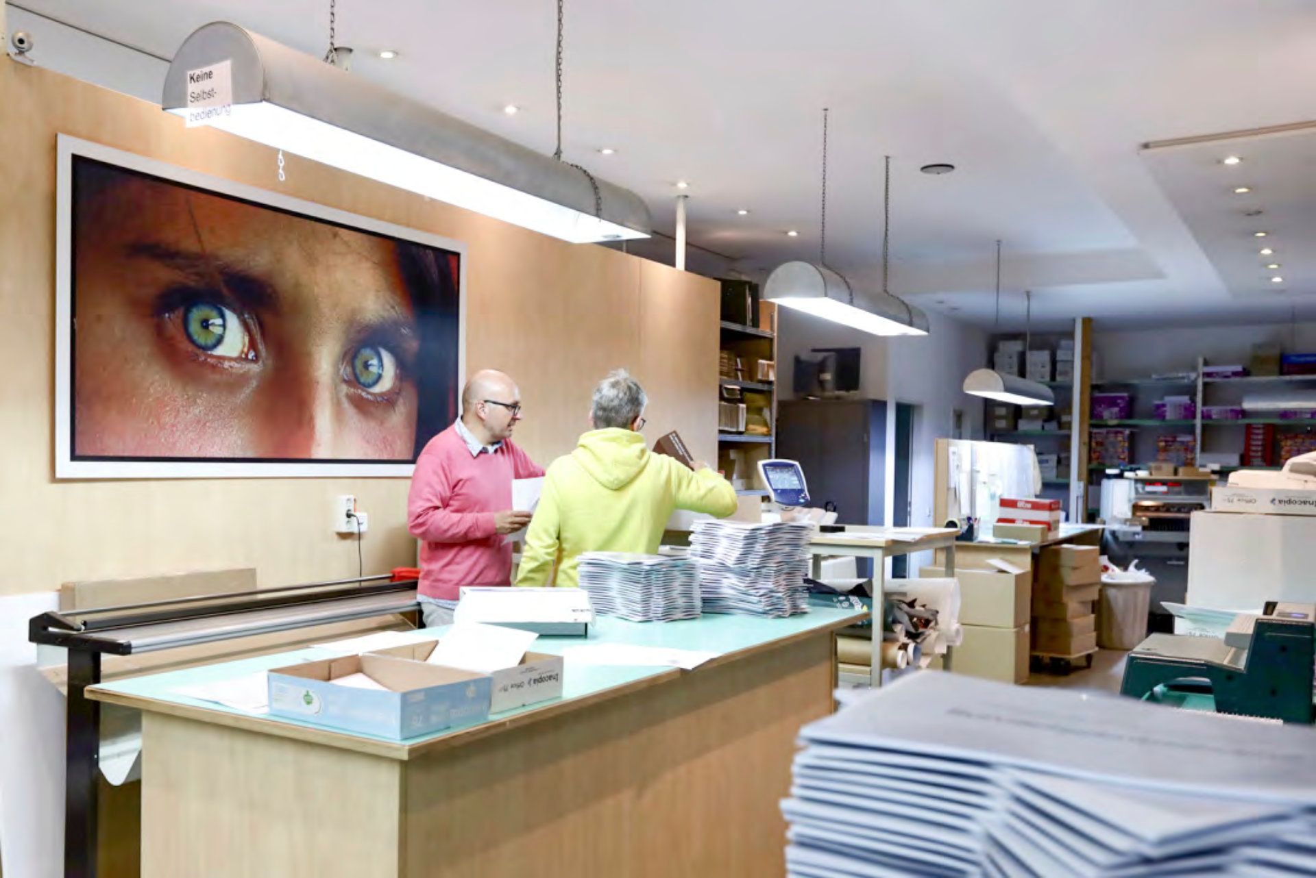 Zwei Männer arbeiten in einem Raum, an dessen Wand ein großes Bild mit den Augen einer Frau hängt.