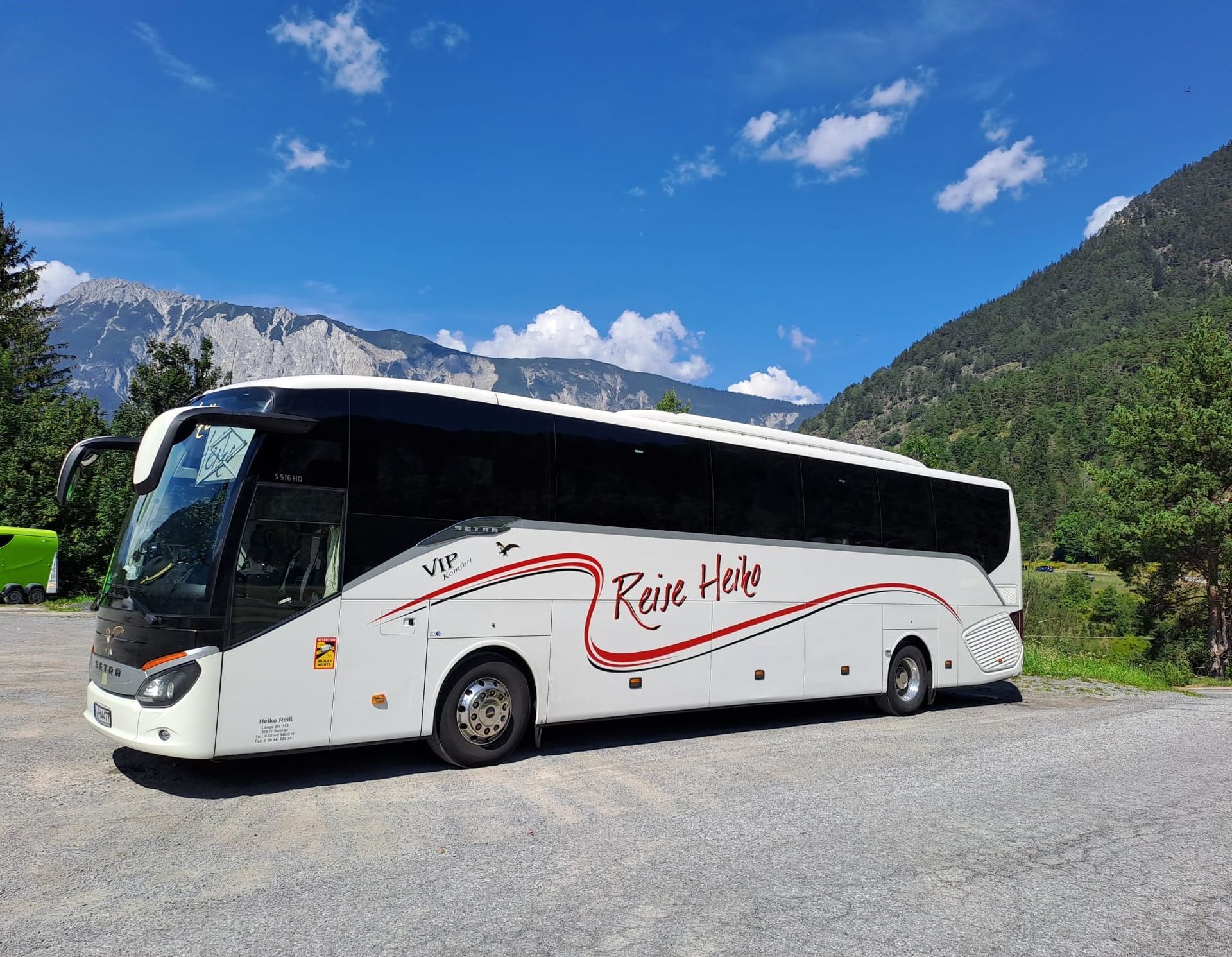 Ein weißer Reisebus parkt auf einem Schotterweg, im Hintergrund Berge und blauer Himmel. An der Seite befindet sich das Logo „Rare Haim“.