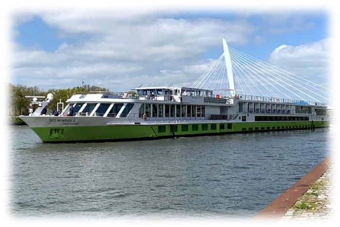 Grün-weißes Flusskreuzfahrtschiff auf dem Wasser, mit einer modernen Brücke im Hintergrund.